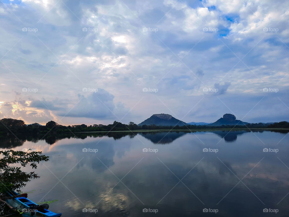 Reflection of Sigiriya rock fortress and Pidurangala rock visible on a nearby lake. Morning view with sun rising from a corner. Seeing doubles.
