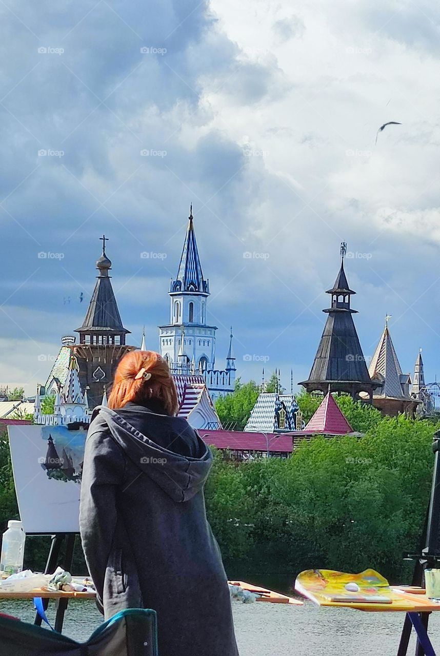 Pond shore. There is an easel on the shore, on which a part of the landscape is painted.A woman stands nearby and draws a landscape.On the opposite side are the dome of the stave church and the domes of the towers among the green trees. Seagulls fly