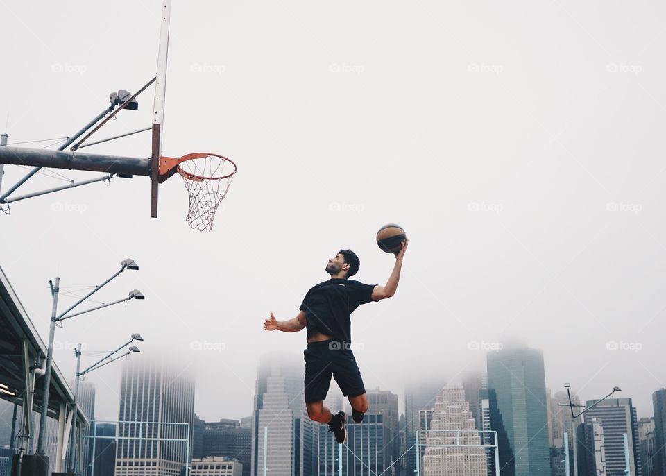 A man playing basketball doing a slam dunk. A city covered in fog is in the background.