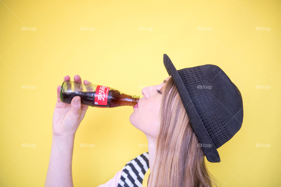 Portrait of a young woman drinking a bottle of Coca-cola in front of a bright yellow wall