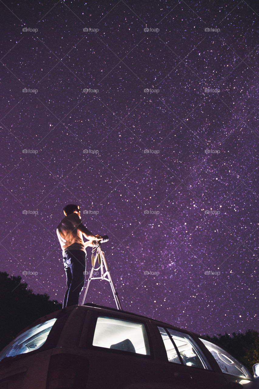 A photographer stands on top of his car to photograph the milkyway stars this is a mix of the natural light from the universe & flashlights to light the photographer up