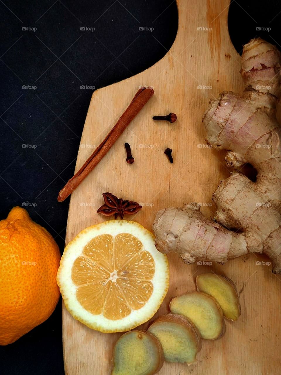 Wooden board containing: sliced lemon, anise, cinnamon stick, ginger root and sliced ginger. There is a lemon near the board. Black background