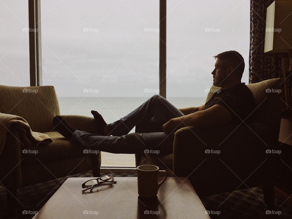 Man relaxing and contemplating while watching the ocean through large window with coffee and glasses