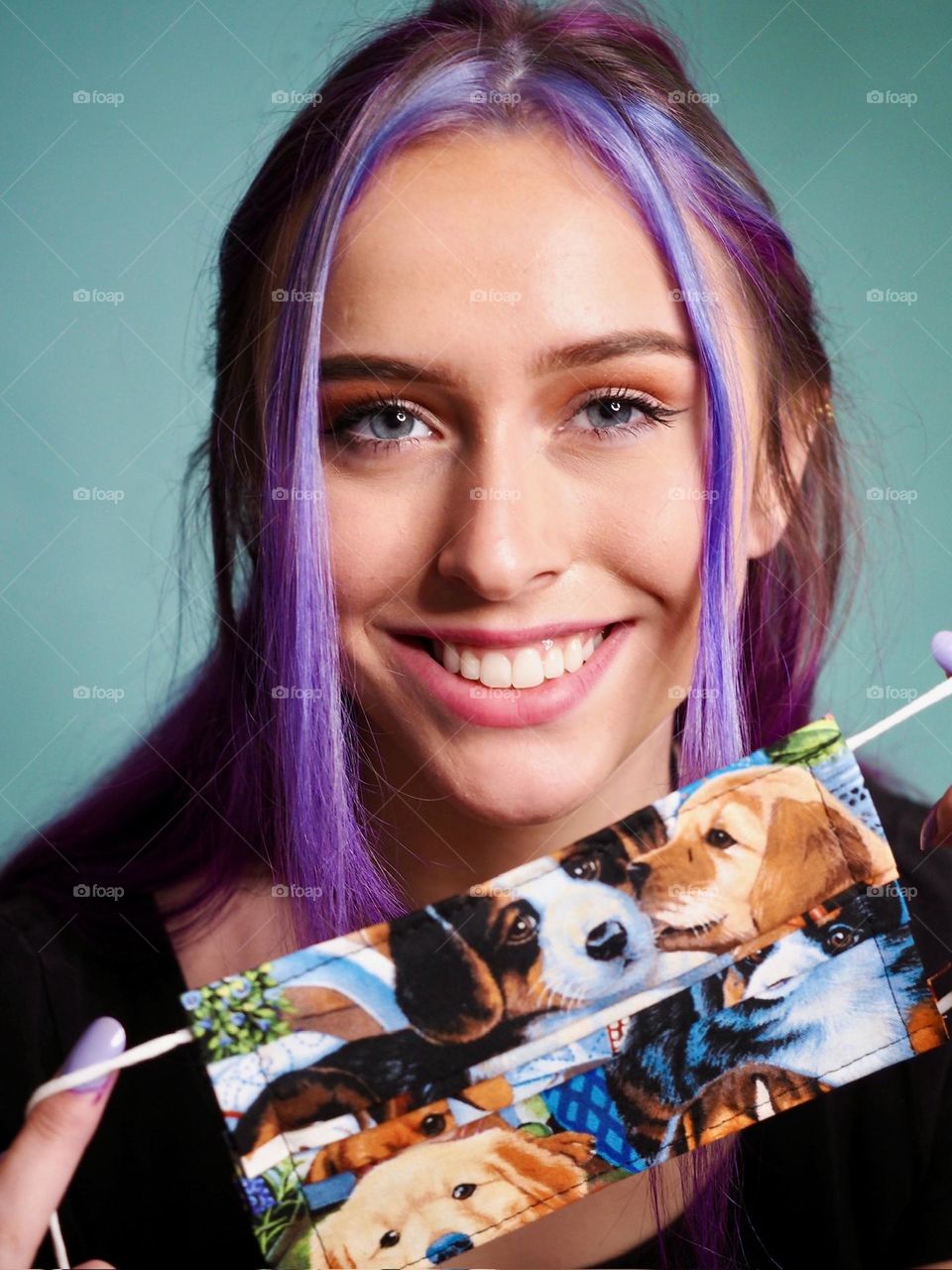 Beautiful young woman with vibrant, violet hair and gorgeous smile, holding handmade face mask featuring puppies, studio portrait
