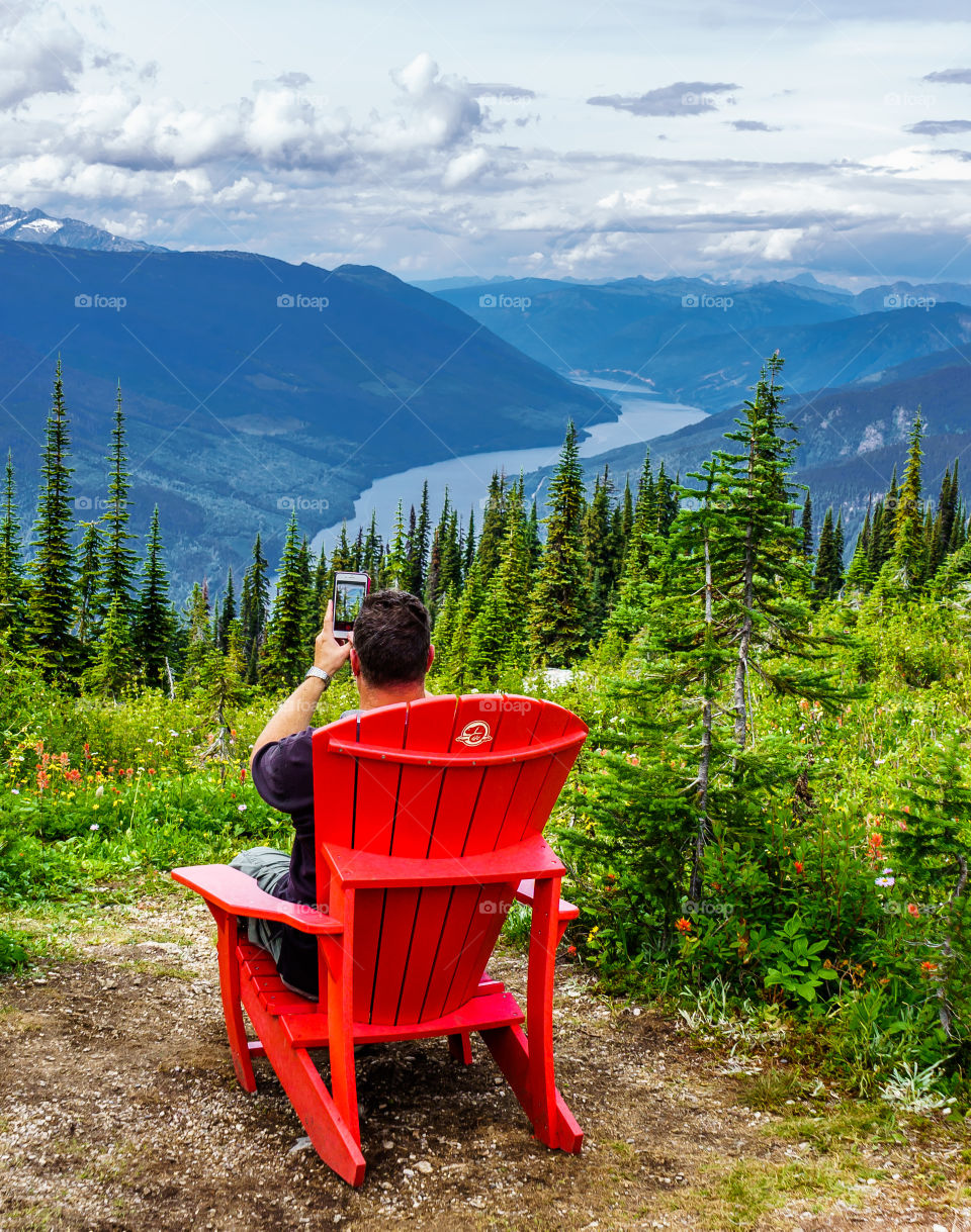 Man relaxing on top of Mount Revelstoke and taking a picture of the scenic surroundings - overlooking Columbia River valley