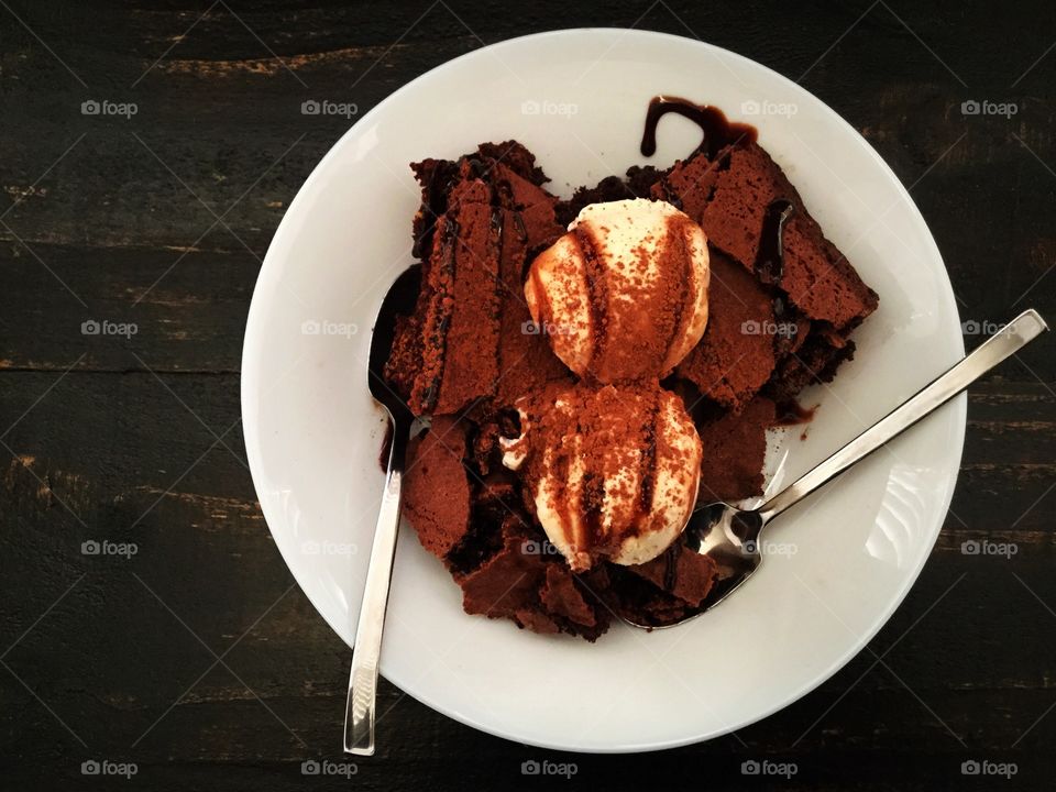 Brownies topped with vanilla ice cream, chocolate sauce, and cocoa powder and shown with two spoons
