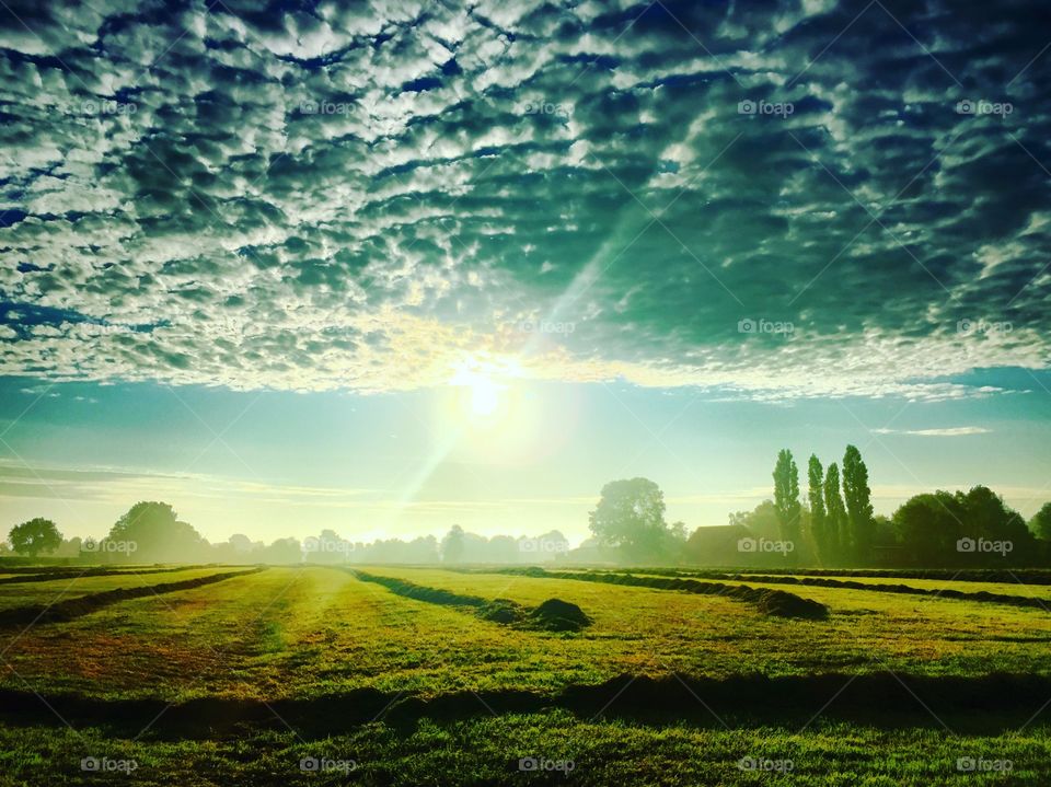 Sun sneaking from under a thick layer of clouds over a farming grassland which if recently mown