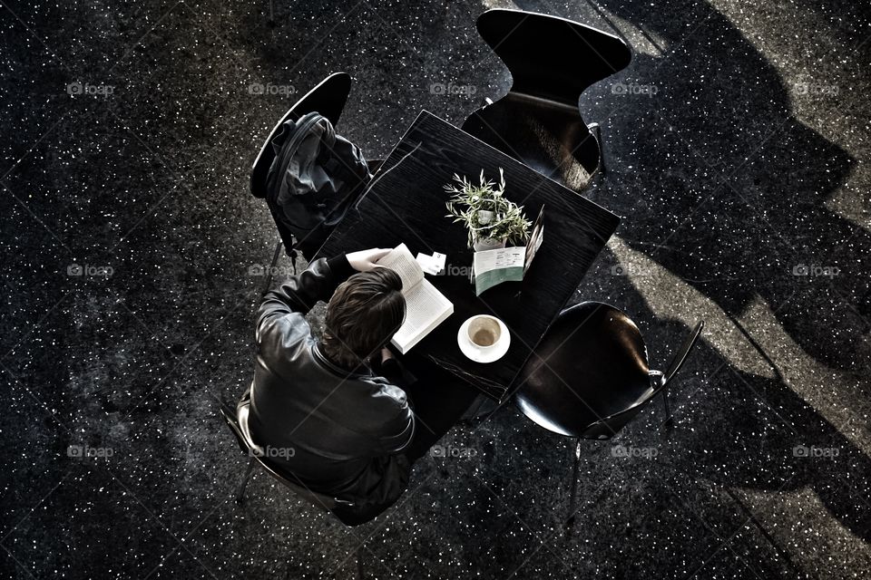 Overhead shot taken from a gallery balcony of a guy reading a book with a cup of coffee ... light streaming in from huge floor to ceiling windows.