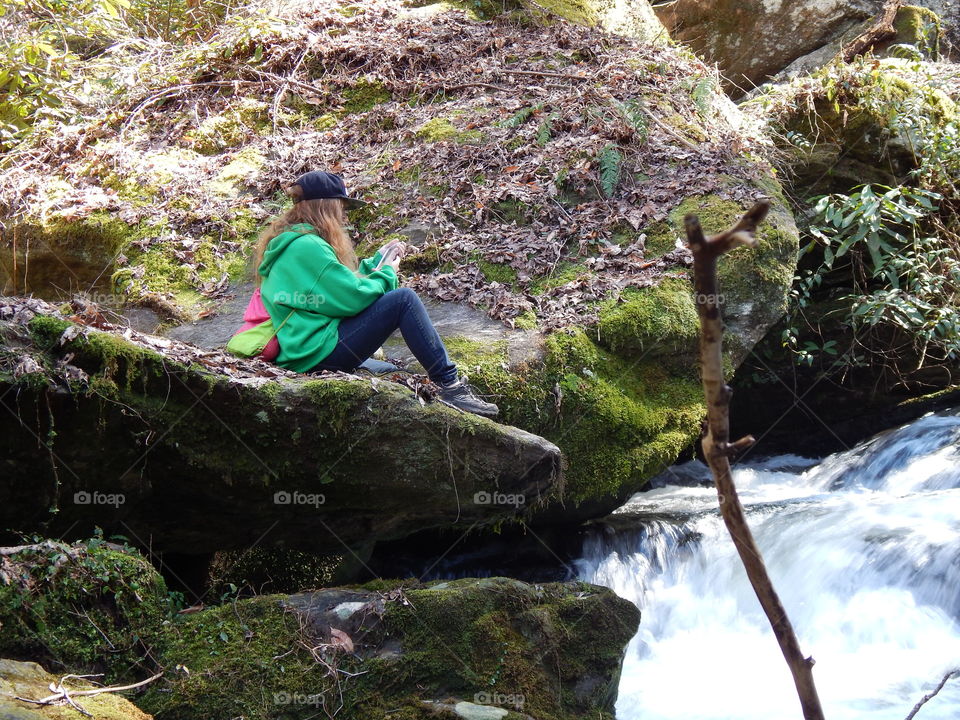 young girl setting on giant moss covered boulder admiring a roaring mountain waterfall