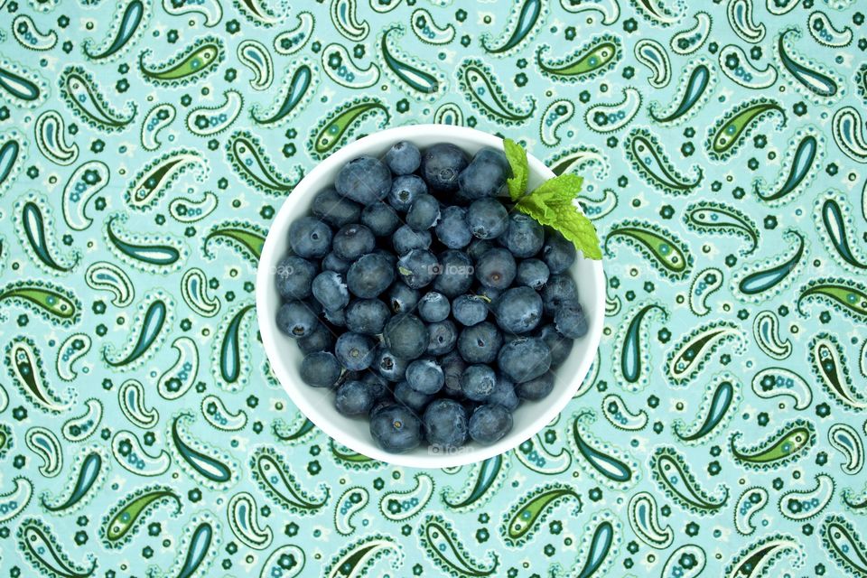 Blueberries and a spring of mint in a white bowl on teal and green paisley fabric