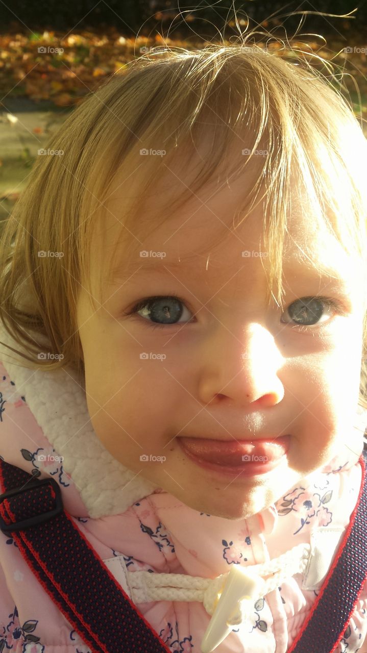 close up of face of young blue eyed blonde haired toddler smiling poking out toungue.