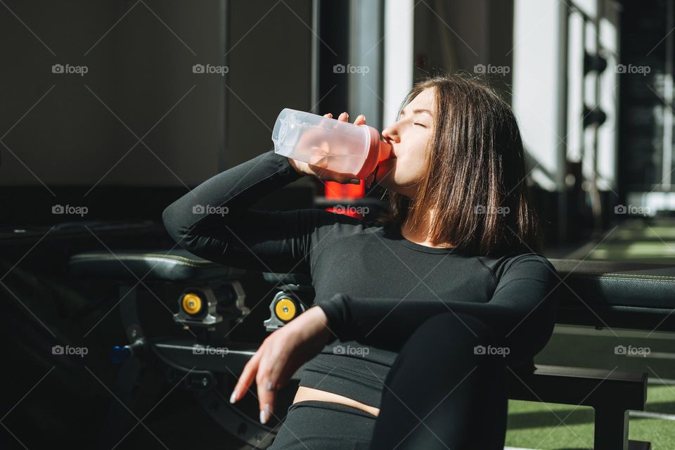 Portrait of resting young brunette woman in sport active wear drinking water in the fitness club gym