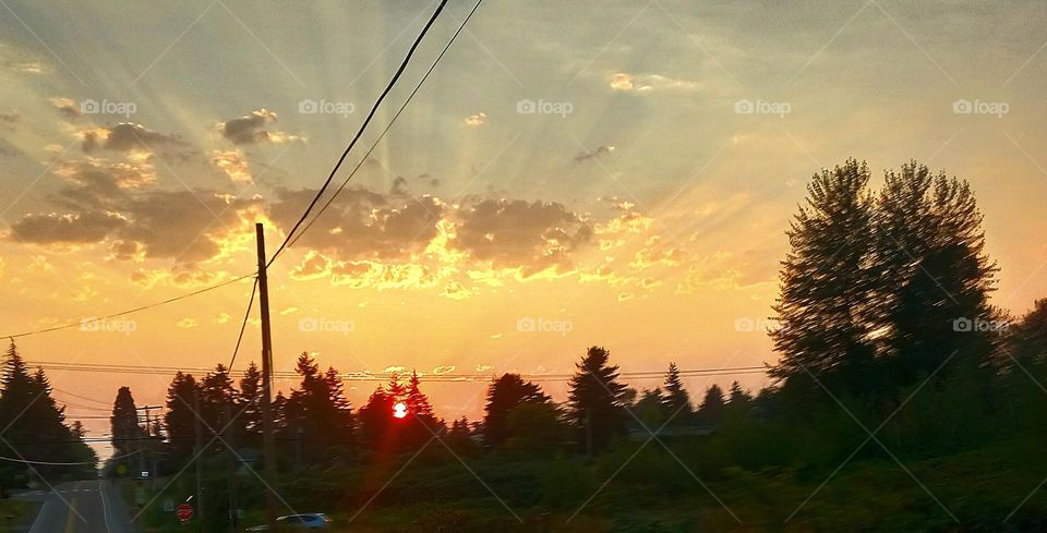 A vivid display with a bright reddish sun orb, slipping over the tree lined hill with white and gray clouds shooting sun rays over the city streets.