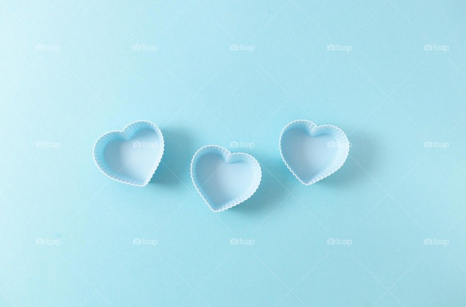 Three light blue heart-shaped muffin cups lie in a row in the center on a soft background, flat lay close-up. Concept blue on blue.