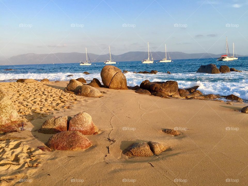 Golden sandy beach with rocks, foamy sea waves and four white sailboats in the background, Porto Apollo, Corse in the background 