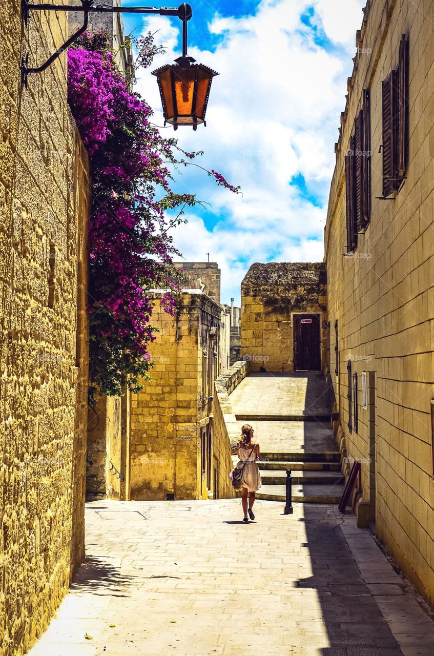 A woman walks the historic streets of Mdina in Malta as purple Bougainvillea blossom on walls