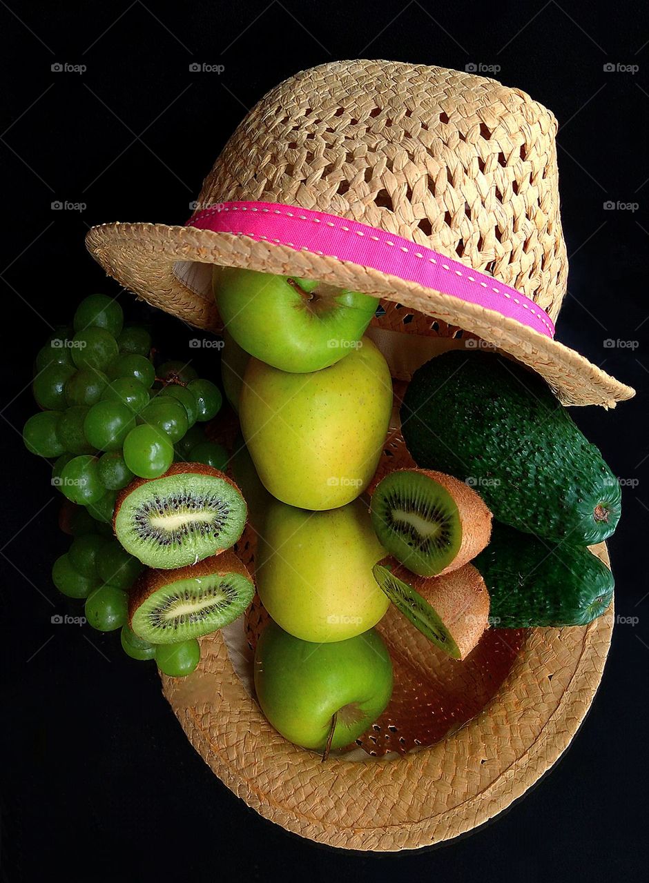 Green color.  The background is mirror black.  Composition.  Three green apples, a green grape, a green avocado, and two green kiwi halves lie under a straw hat.  All fruits and the hat are reflected in the mirror surface