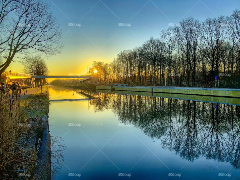 Golden and Blue sunrise or sunset sky reflected in the water of the river or canal which is lined by bare trees and their reflection in the water