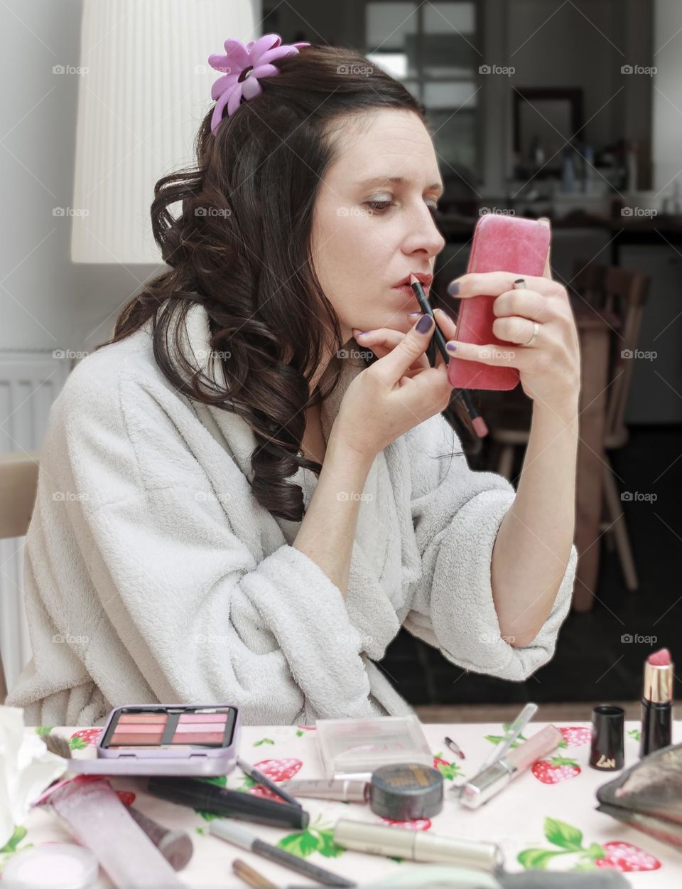 A woman sits at a table covered in make up as she applies lip liner while looking into a small, well used hand mirror