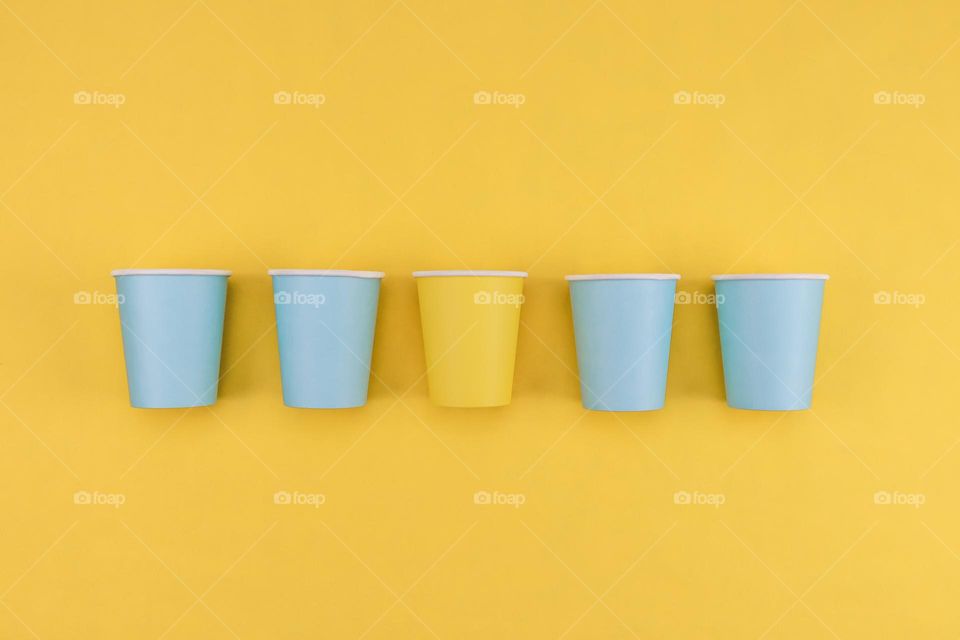 Five paper cups: four blue on the sides and one yellow in the middle lie in a row on a yellow background, flat lay close-up.