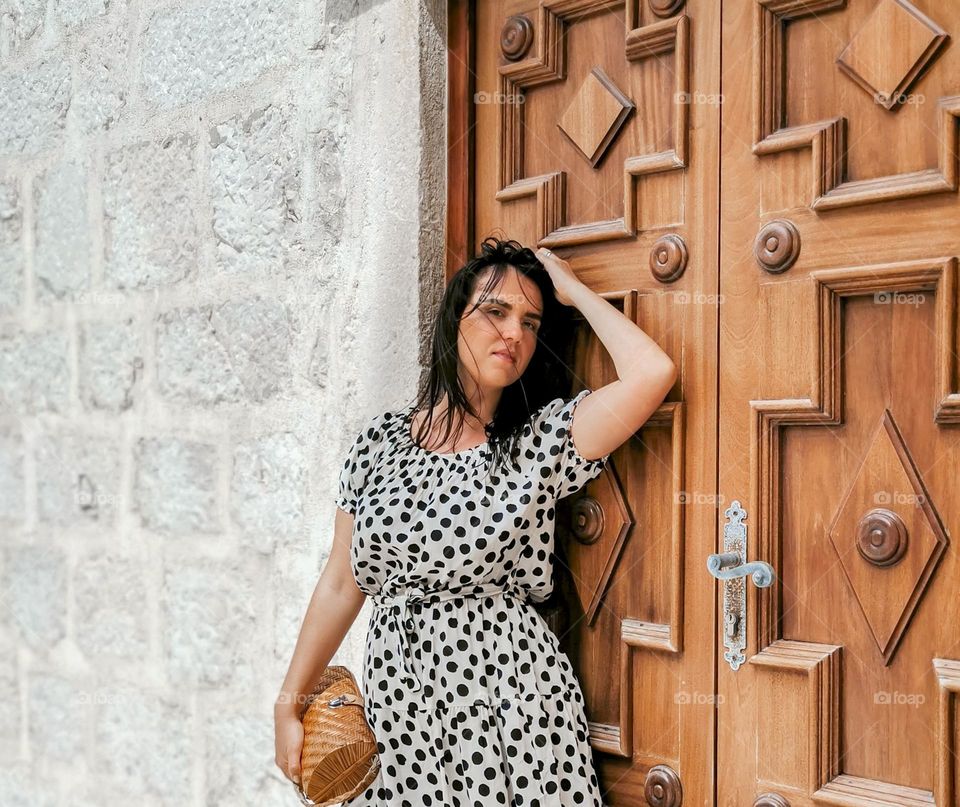 Portrait of beautiful young woman wearing polka-dot sundress, standing in front of wooden door of stone building