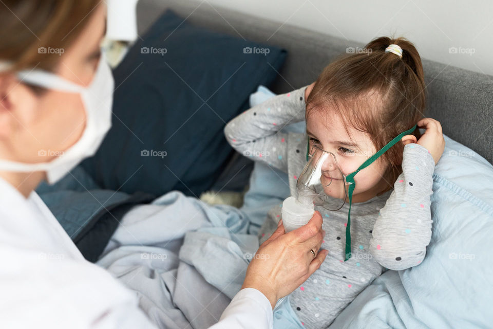 Doctor visiting little patient at home. Child having medical inhalation treatment with nebuliser. Woman wearing uniform and face mask