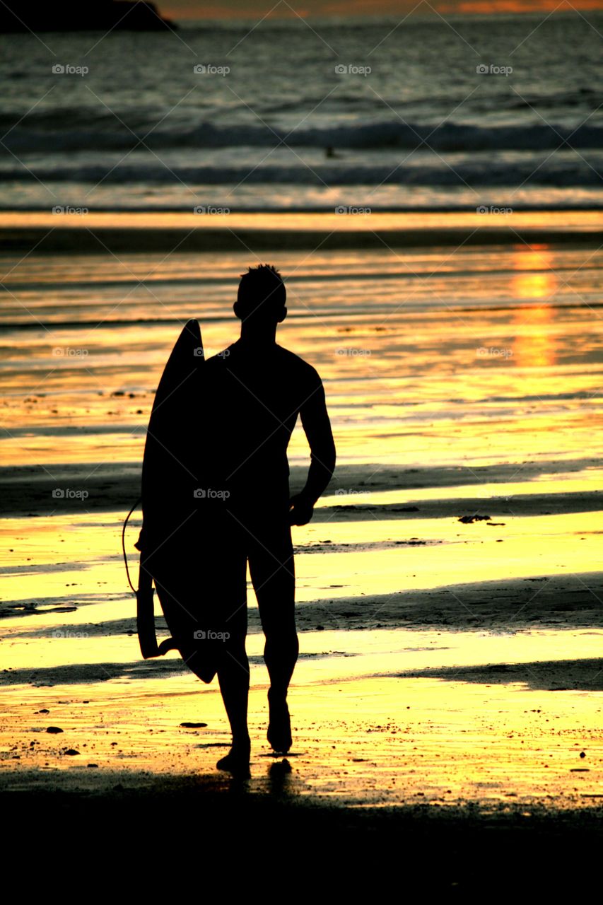 Surfer coming home at sunset. A surfer after a late night surf on Fistral Beach in Cornwall,silhouetted in the wet sand reflecting the sunset 