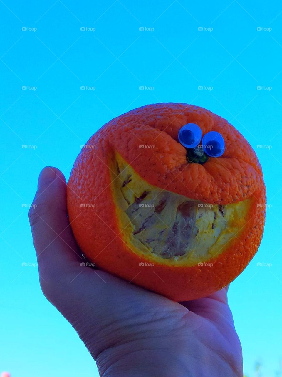 Color contrast: orange and blue. Hand holds an orange orange that smiles. Blue sky in the background.Food. Fruits