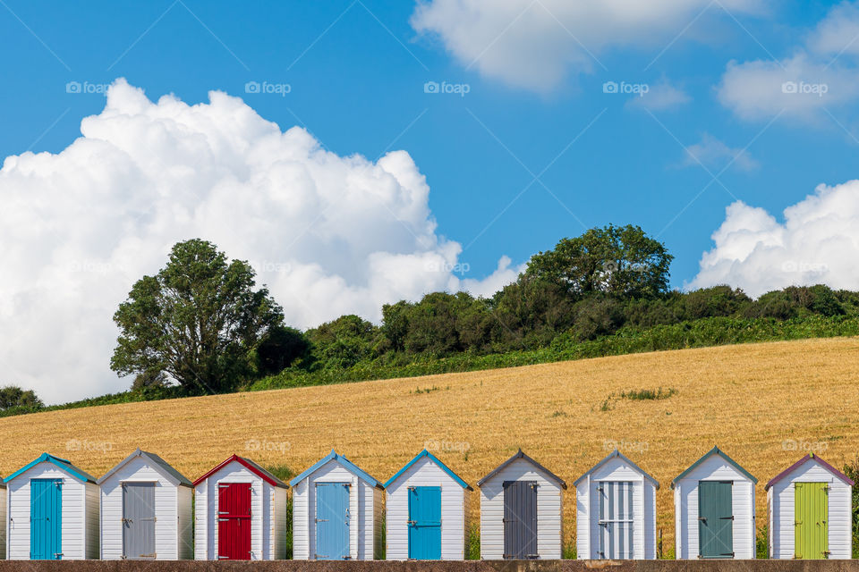 Little colorful beach sheds against fields with treeline and cloudy blue skies.
