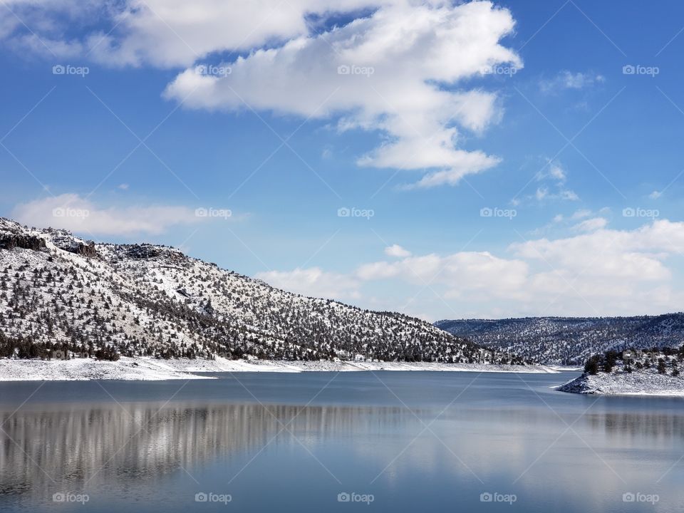 Snow covered hills, trees, clouds, and a bright blue sky reflect in the partially iced over Prineville Reservoir on a sunny winter day in Central Oregon. 