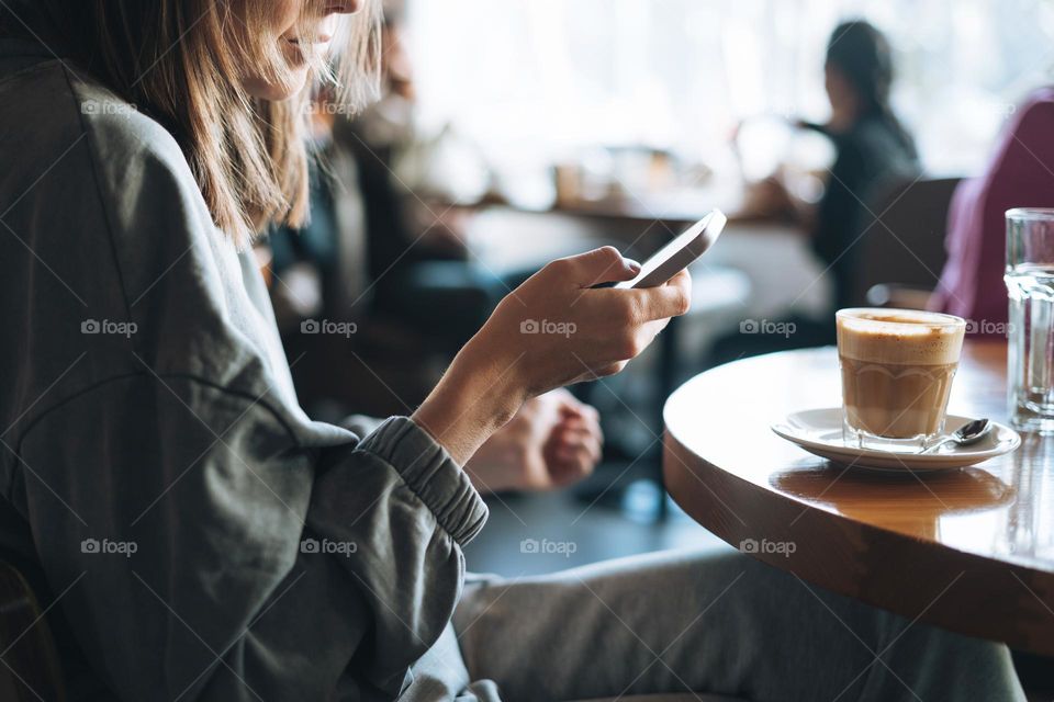 Young brunette woman wearing casual longsleeve with mobile phone in hands with cup of coffee sitting in cafe, city life