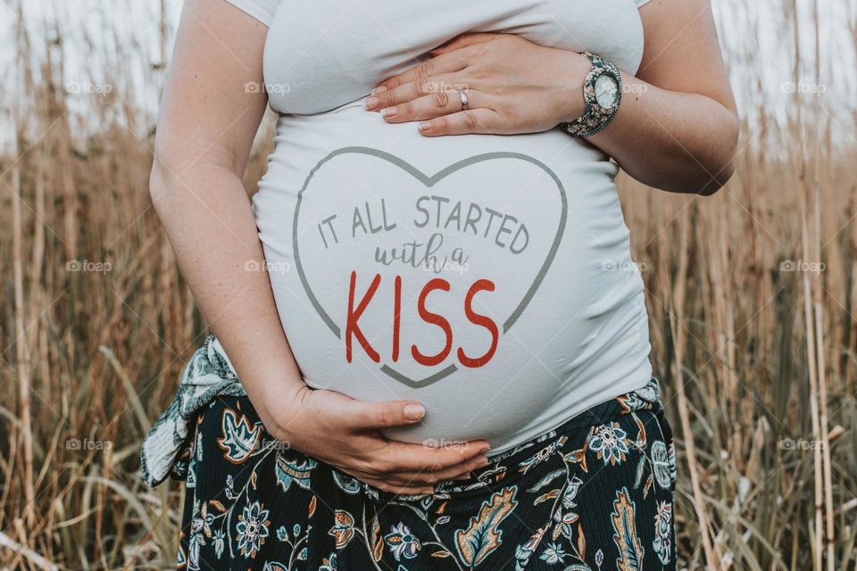 Pregnant woman holding hands on her belly while wearing a funny t-shirt with a writing "it all started with a kiss"