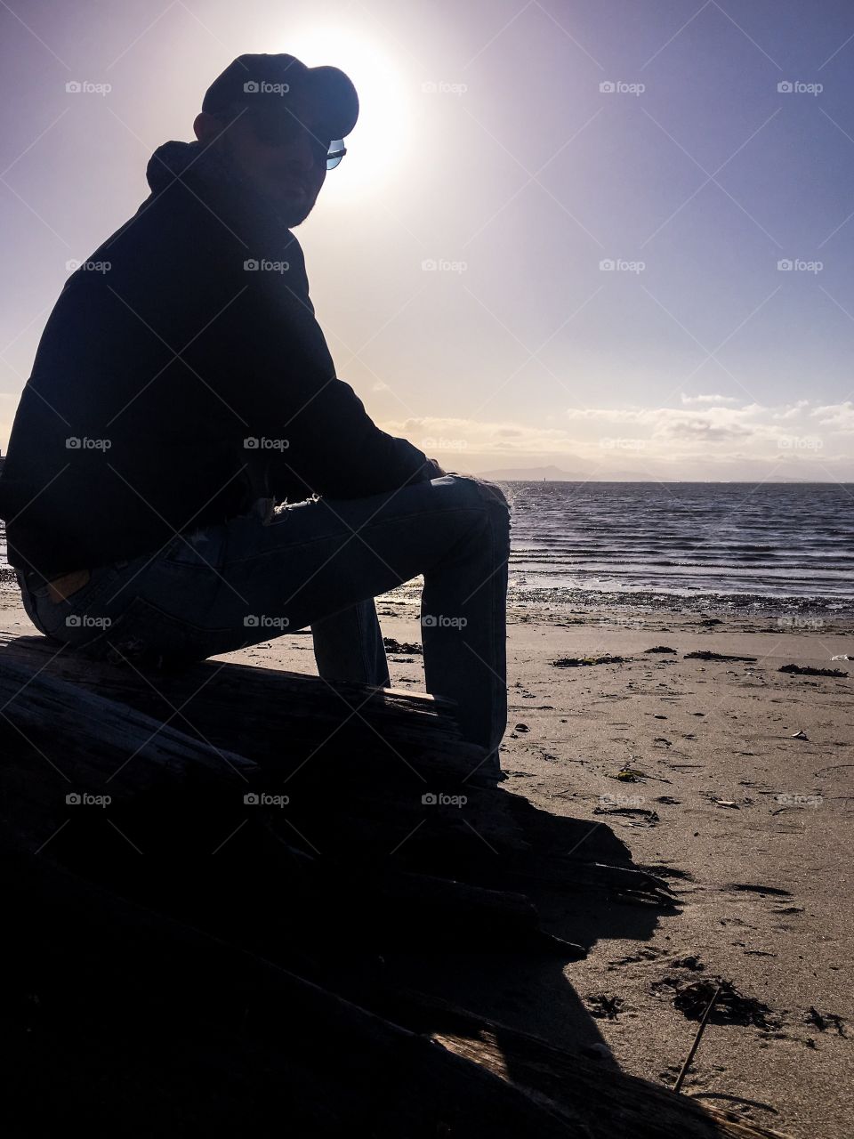 Man sitting on driftwood looking at camera with the sun behind his head.