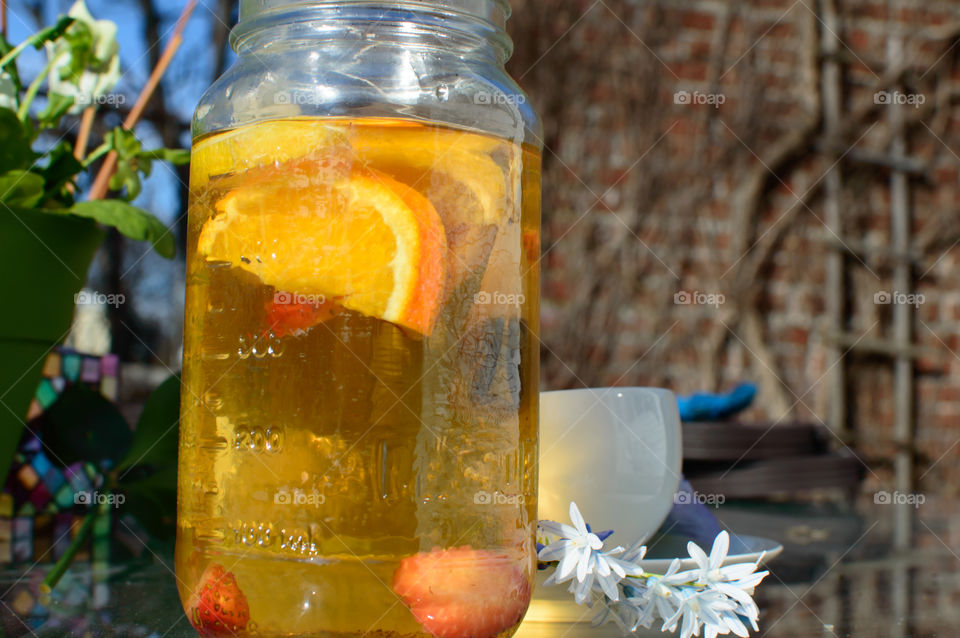 Making steeping fresh herbal tea in glass jar with citrus and fruit using heat of the sun outdoors on patio table with tea cup and flowers with blue sky and brick wall background