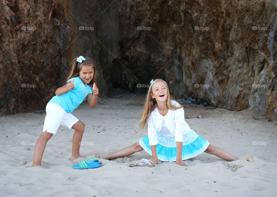 Two young girls dressed in coordinated teal blue and white outfits playing on beach acting funny and silly. Genuine and authentic energy, joy, happiness and smiles. Long hair, shorts, skirt, hair accessories 