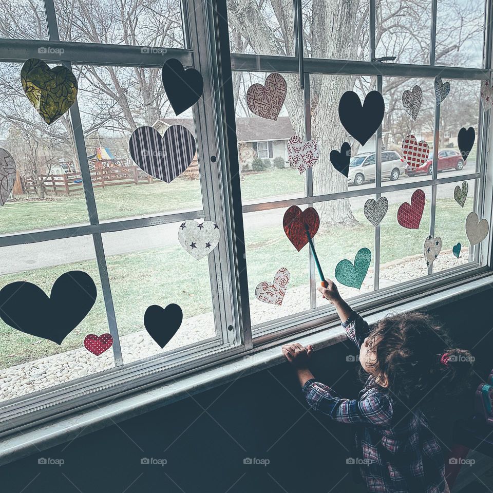 Toddler girl pointing to hearts on window, toddler peering out window, child looks outside through window panes, child reaching for the decorations, child at the window, toddler enjoying the Valentine’s Day decor on windows, toddler in the window 