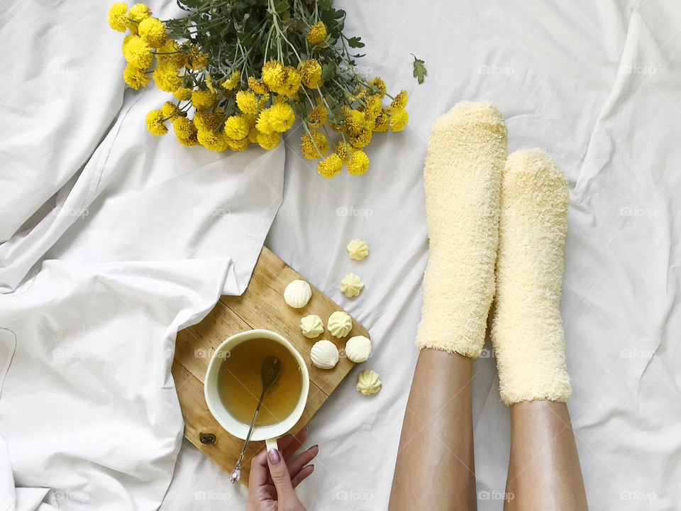 Young woman in cozy yellow socks drinking tea with yellow candies at cozy bed 