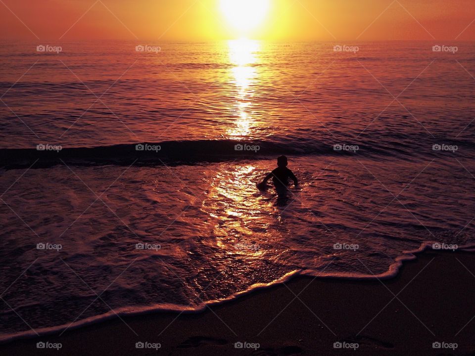 Silhouette of a small boy playing in the ocean surrounded by an orange and gold sunset.