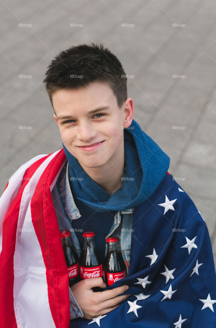 handsome guy, teenager, wrapped in an American flag, sitting on a bench, smiling and holding a Coca-Cola