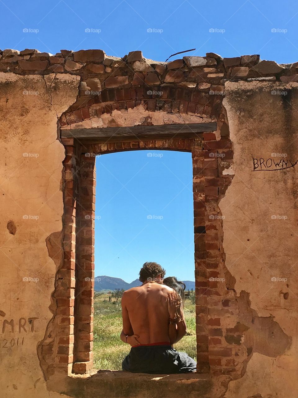 Young couple kissing and embracing in brick doorway of old brick house ruin in rural outback South Australia, with view of Flinders Ranges and outback bush, sunny blue sky viewable through the portal 