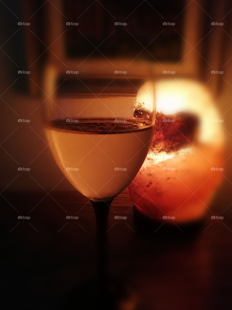 A see-through wine glass with a black leg standing on the brown table in a dark room. The surface of the glass is wet outside because of the cold drink.Some clear drops become visible.Behind is an orange light,made of salt and a painting on the wall