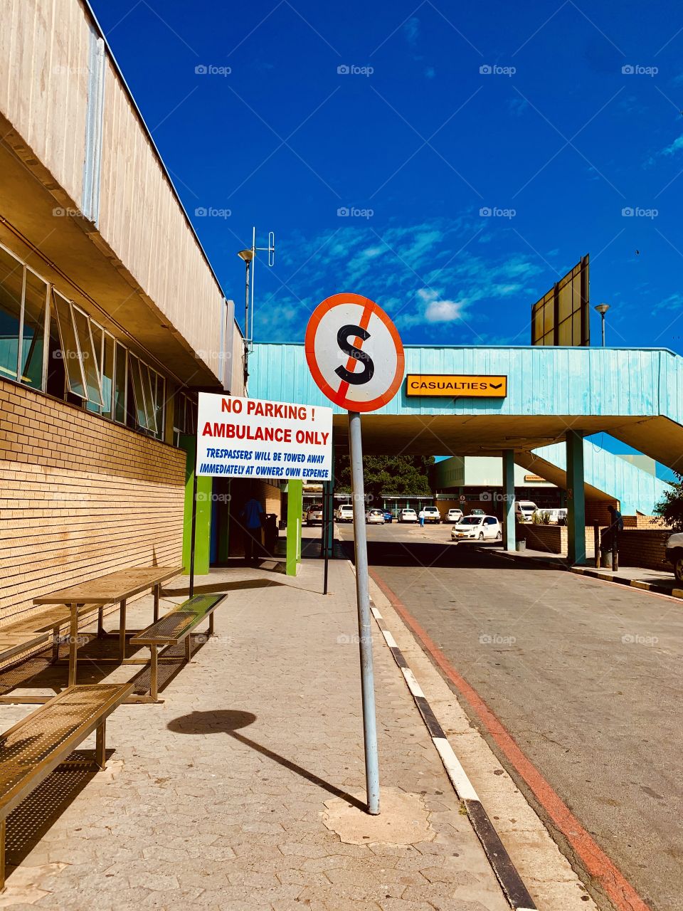 Road signs along the drive through road of the emergency department at the hospital. A no stopping sign for public and private vehicles except for the ambulances, a supplementary sign after that explains the fate of stopping. 