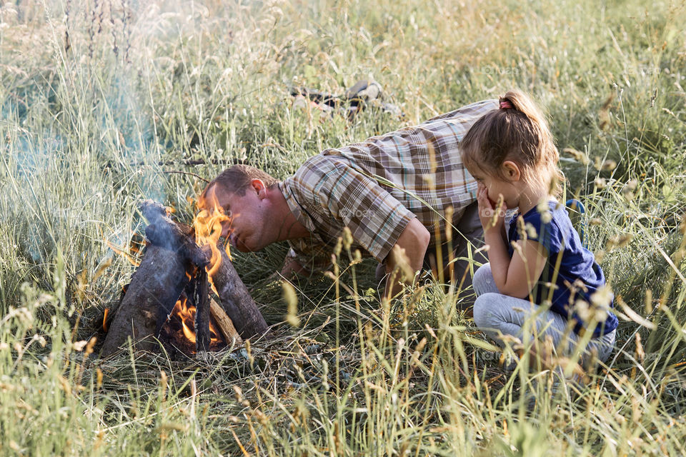 Man starting a campfire, blowing on a fire. Little girl sitting in a grass beside a campfire. Candid people, real moments, authentic situations