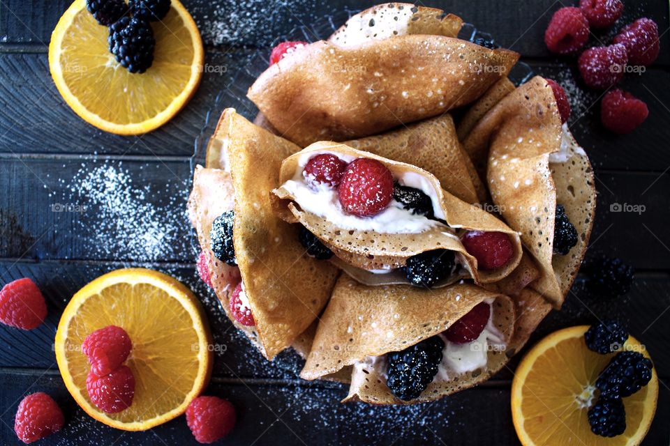 A plate of homemade cream and berry filled crepes on a dark wooden board with orange slices, raspberries and blackberries around it. 