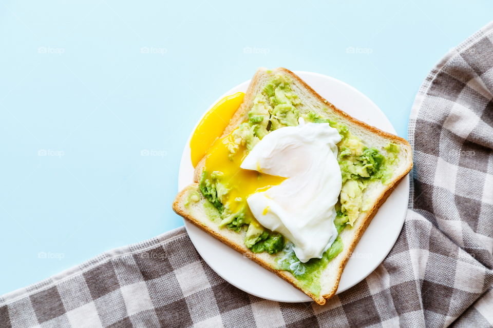 Sandwich with fresh green avocado, poached egg on white plate lying on blue background 