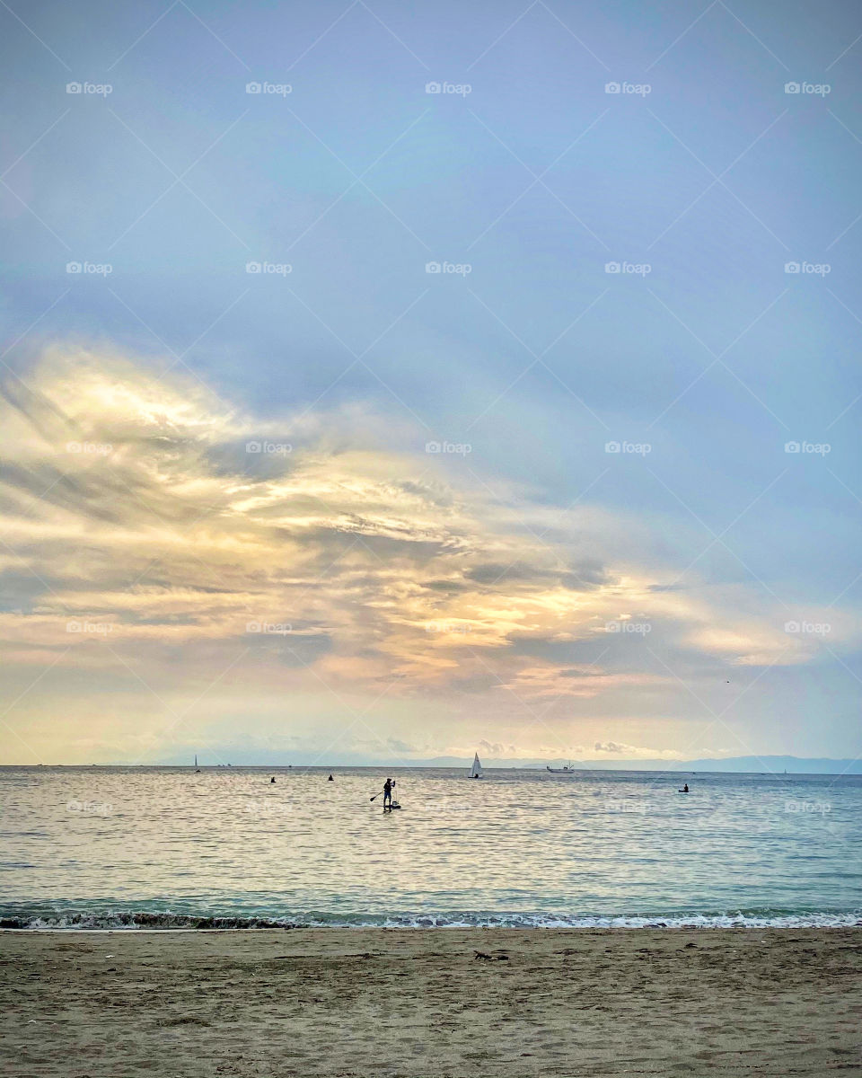 Pastel-colored sea and sky, sunlight turning clouds a muted toned-down orange. SUP and sailing activity going on in this serene ethereal-looking scene.