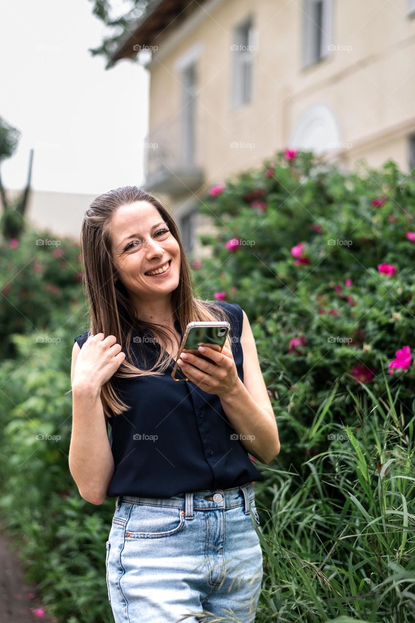 A young girl in a blue blouse and jeans looks at the camera and laughs, holds a phone in her hands, a walk in the park, wild rose blooms in summer, people use modern technologies