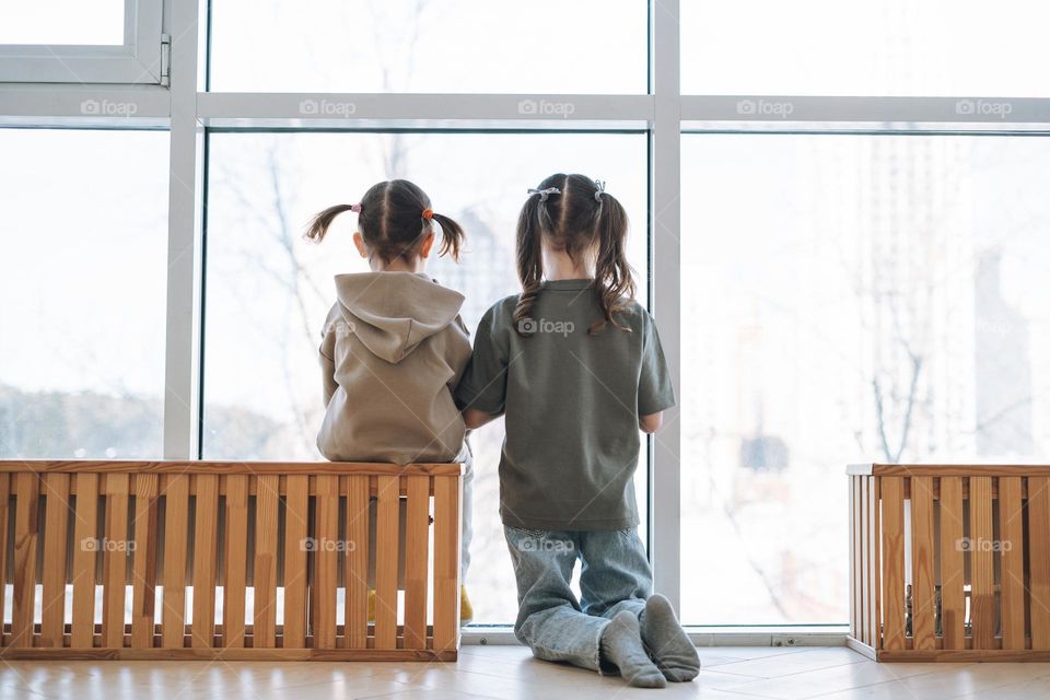 Cute little sisters girls looking in window at home, people from behind