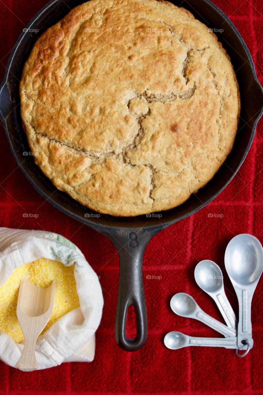 Flat lay of sourdough cornbread in a cast iron skillet, cornmeal and wooden scoop in a flour sack, vintage measuring spoons on a red kitchen towel