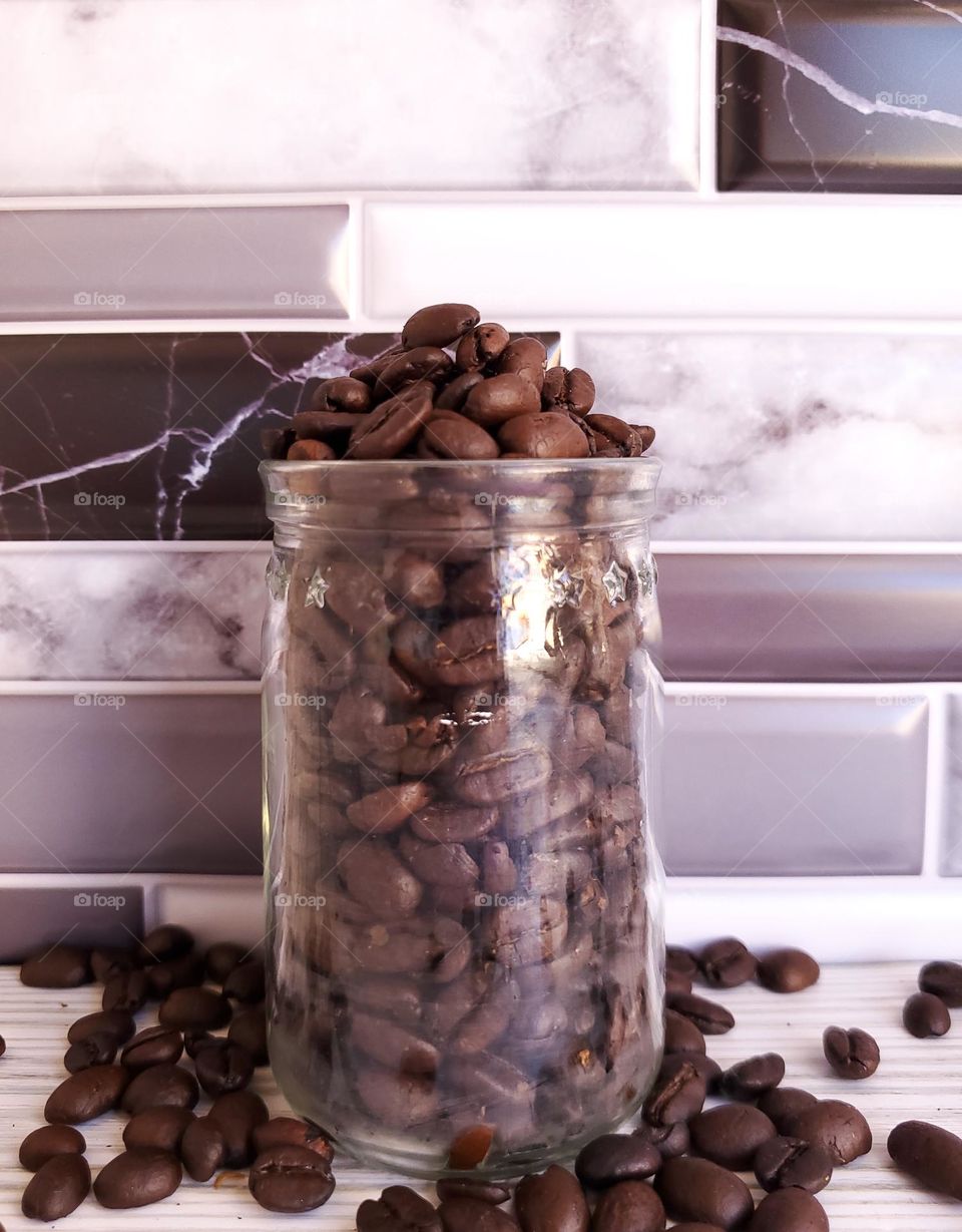 A clear glass jar with overflowing coffee beans in front of a violet gray, white and black tile backsplash.